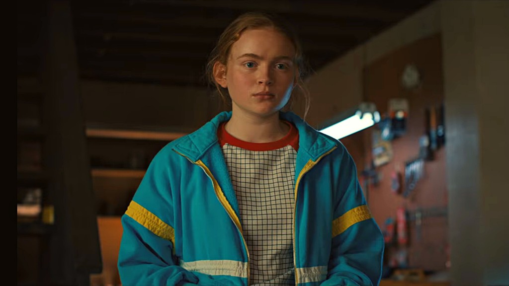 Max Mayfield in blauer Jacke mit gelben Streifen und rotem Kragen, in einer Werkstatt