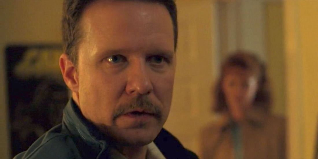 Neil, Schnurrbart und dunkle Jacke, im Flur, Susan unscharf hinter ihm.
